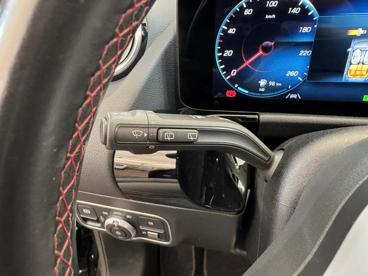 Gros plan sur le volant noir avec surpiqûres rouges et la commande d’essuie-glace, tableau de bord numérique visible en arrière-plan.