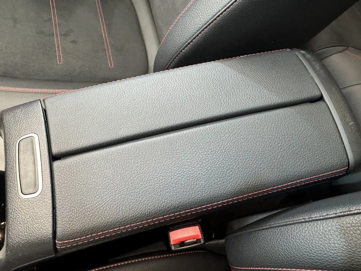 Gros plan sur l'accoudoir central en cuir noir avec surpiqûres rouges, sièges en tissu noir partiellement visibles.