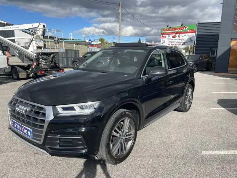 Audi Q5 2.0 TFSI 252 AVUS noir métal vu en 3/4 avant droit sur parking extérieur sous ciel partiellement nuageux