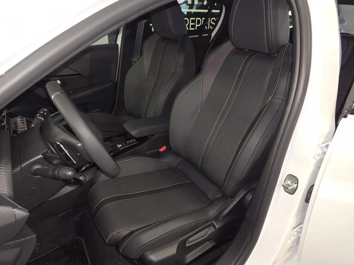 Vue intérieure côté conducteur d’une Peugeot 208 blanche 2025, siège tissu noir avec surpiqûres vertes et volant compact cuir.