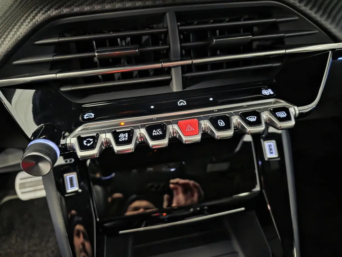 Gros plan sur la console centrale noire brillante du Peugeot 208 1.2i Hybrid 2025 avec boutons métalliques et éclairage LED.