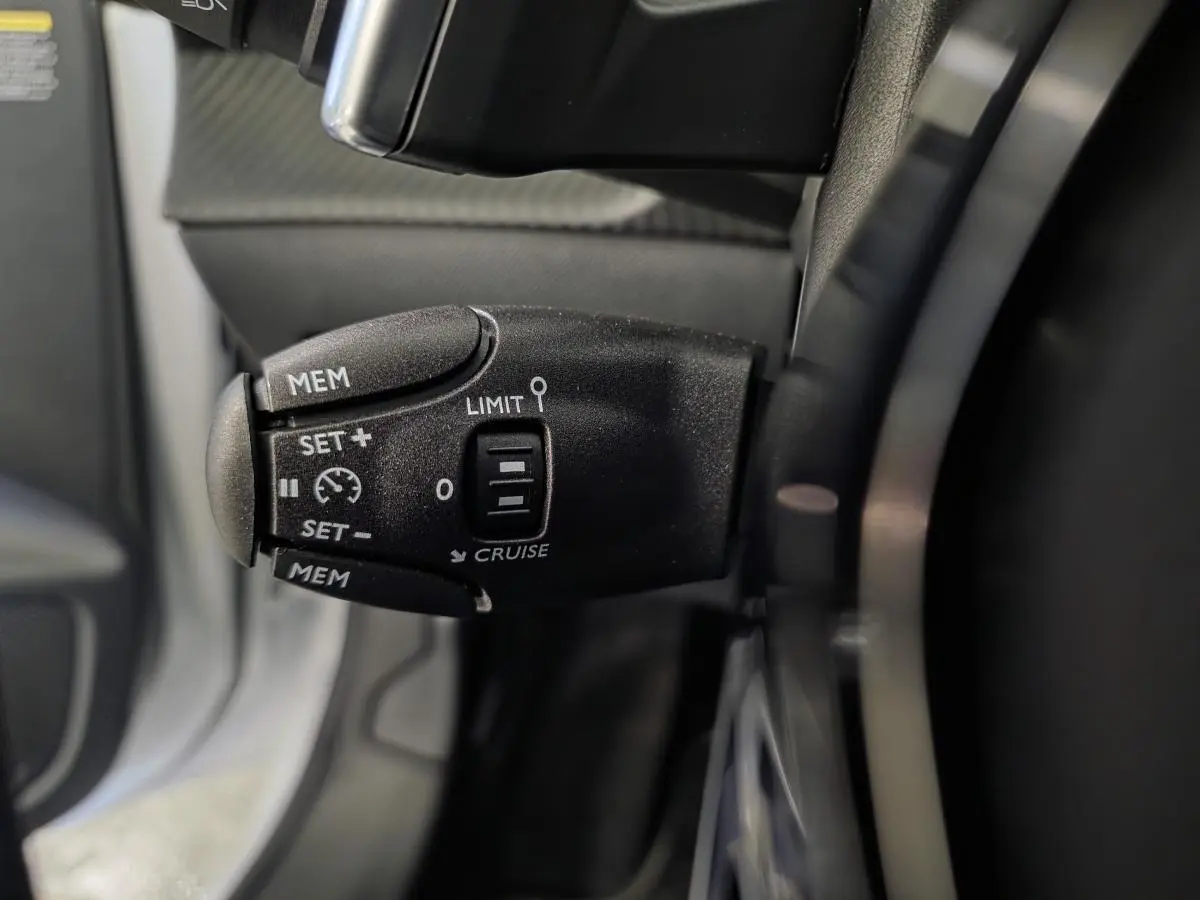 Gros plan sur la commande de régulateur de vitesse noire à gauche du volant dans l’habitacle du Peugeot 208 blanc Banquise.