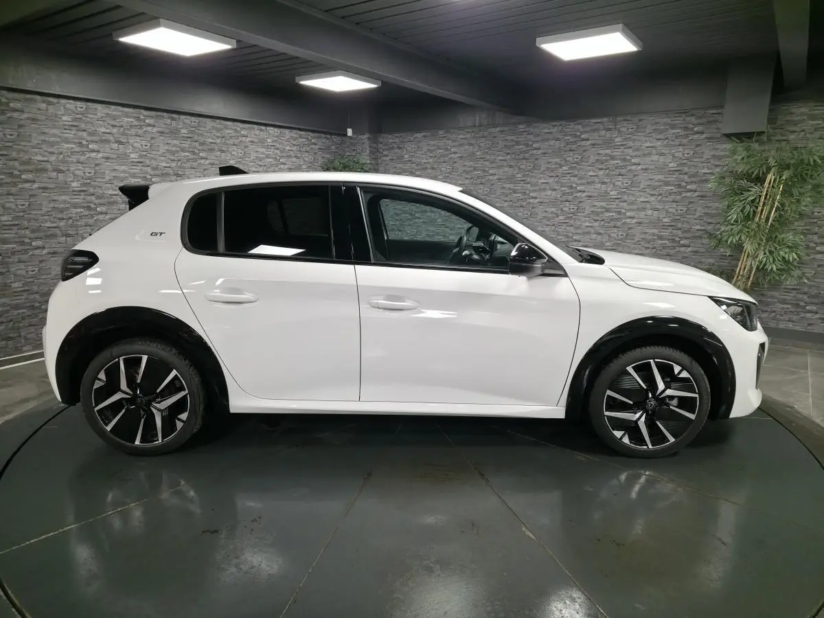 Profil droit d'une Peugeot 208 blanche 2025 GT avec jantes alliage bi-tons et élargisseurs d'ailes noirs brillants.