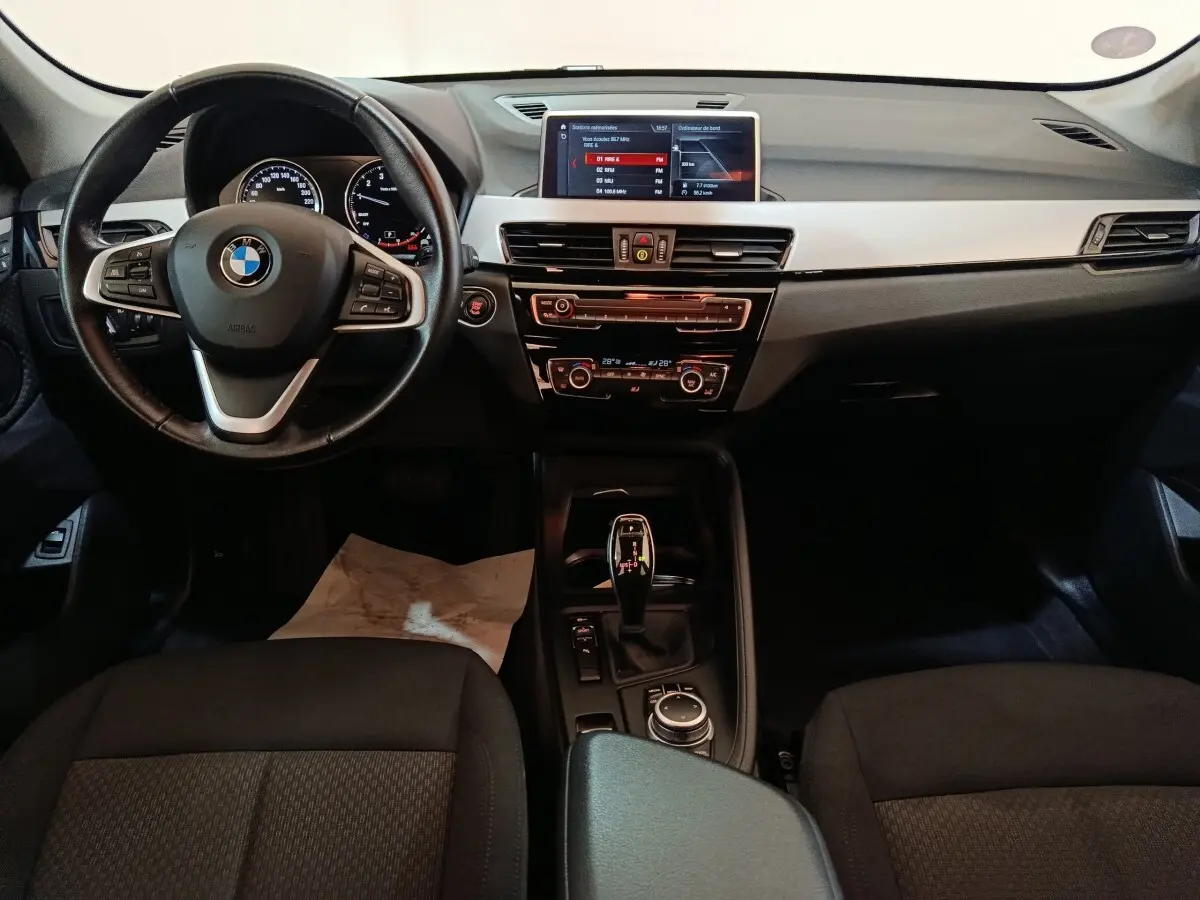 Vue intérieure avant de la BMW X1 sDrive18i 2021, tableau de bord noir avec écran central et volant cuir multifonctions.