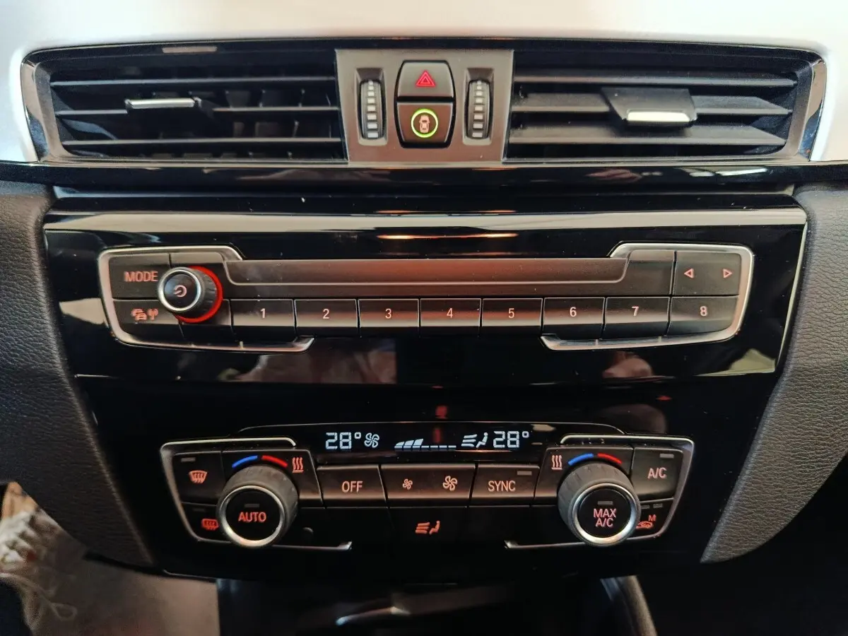 Vue rapprochée du tableau de bord noir brillant de la BMW X1 sDrive18i Lounge avec commandes climatisation digitale à 28°C.