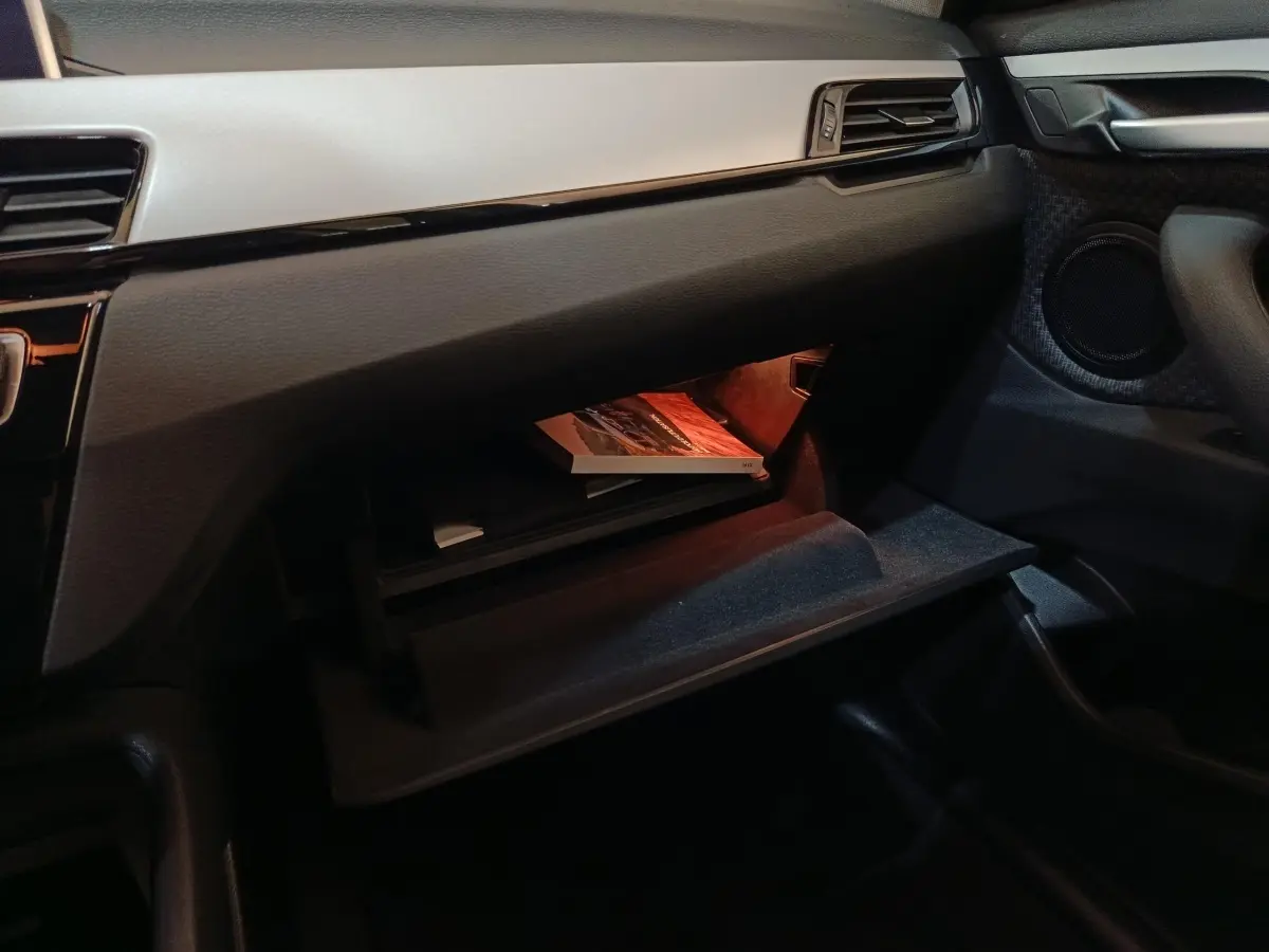 Intérieur BMW X1 sDrive18i 2021, vue côté passager sur boîte à gants ouverte avec éclairage orange et tableau de bord gris foncé.