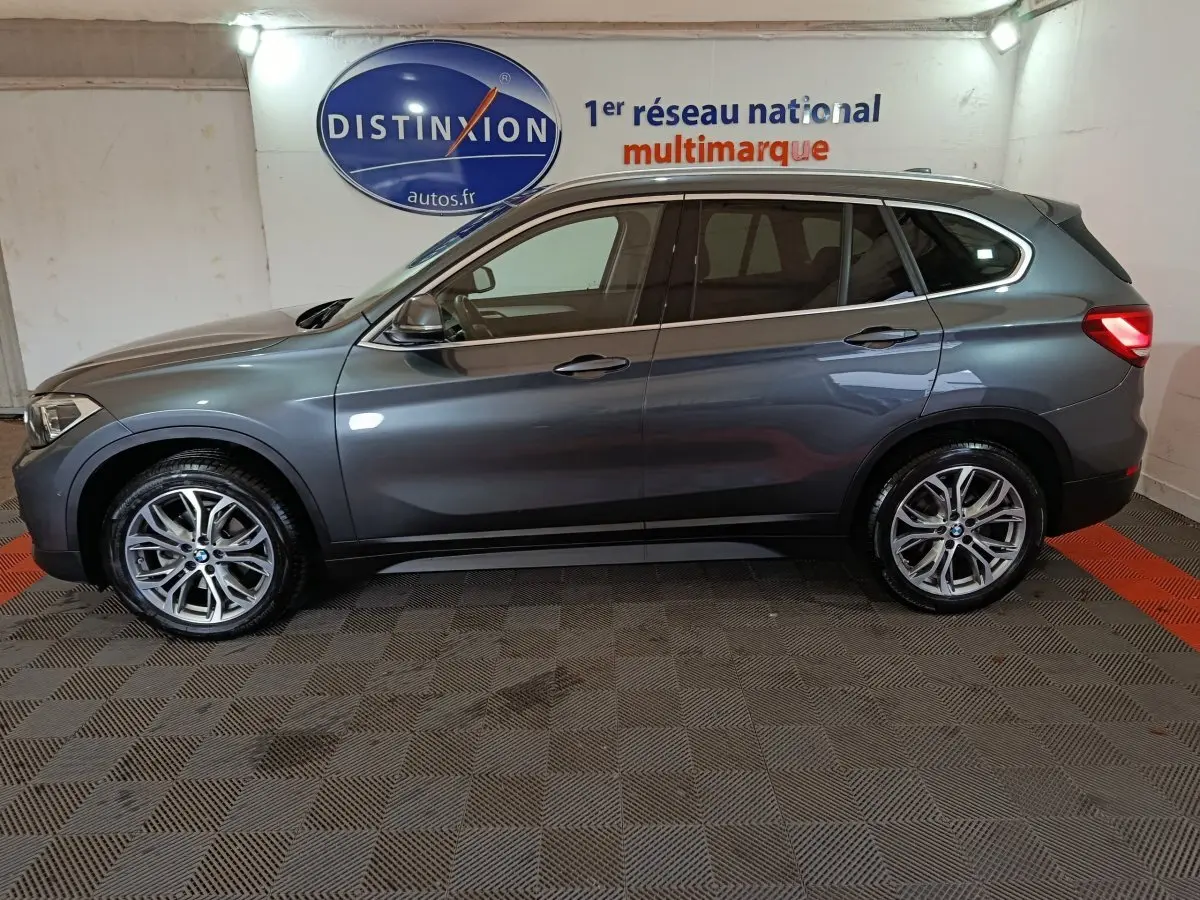 BMW X1 sDrive18i Lounge gris foncé vue de profil côté gauche dans un garage avec logo Distinxion en arrière-plan.