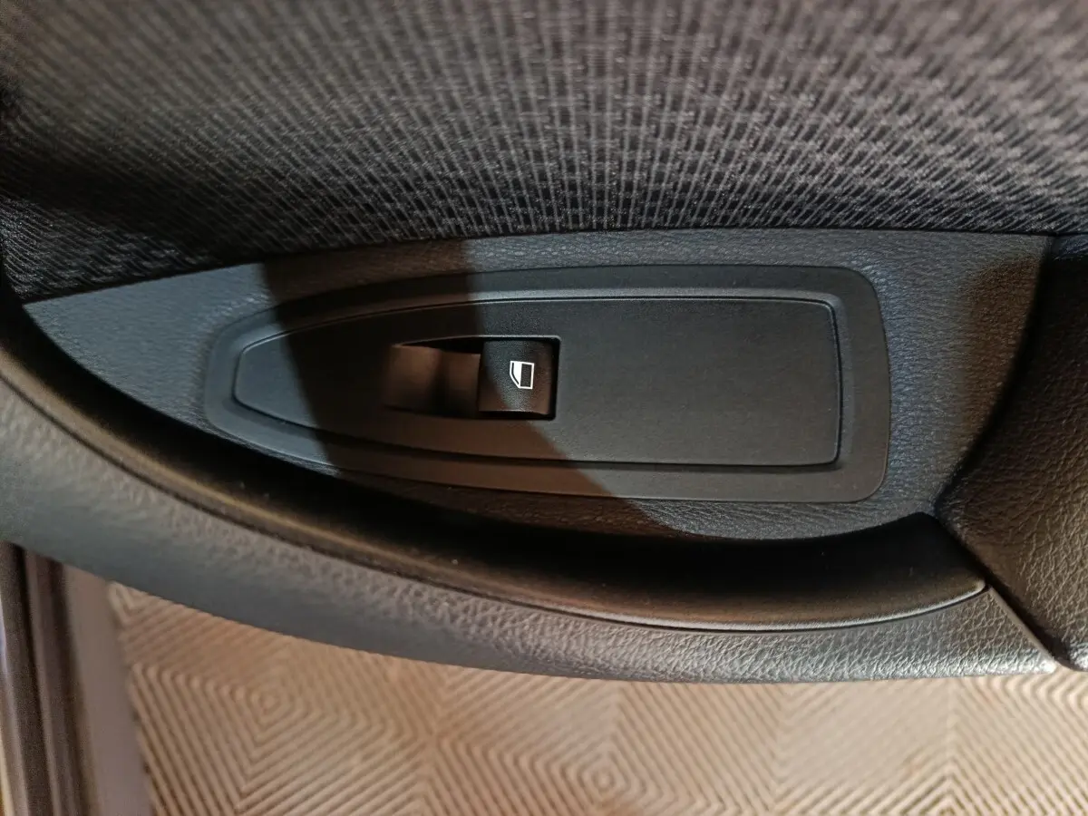 Gros plan sur le bouton de commande de lève-vitre noir dans l'accoudoir intérieur d'une BMW X1 gris foncé.