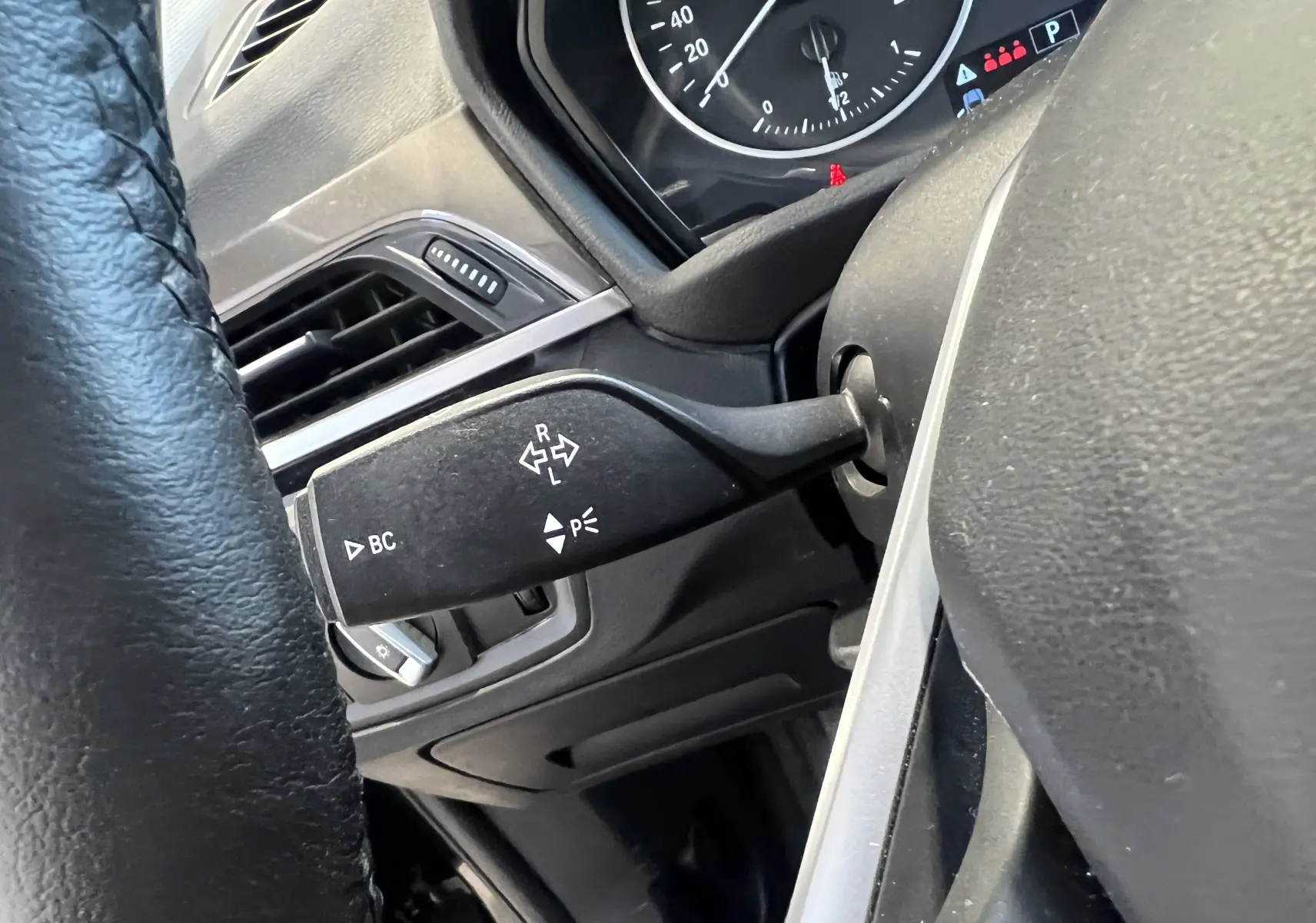 Gros plan sur la manette de clignotant à gauche du volant d’un BMW X1 F48 noir, tableau de bord partiellement visible.
