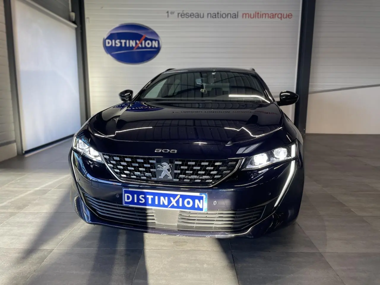 Peugeot 508 SW bleu foncé vue de face en intérieur, avec calandre distinctive et phares allumés.
