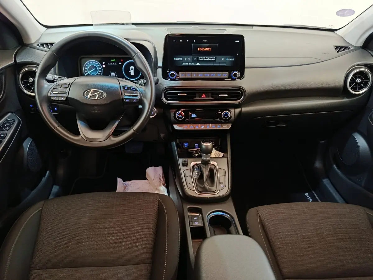 Vue intérieure avant du Hyundai Kona 1.6 Hybrid 2023, tableau de bord noir avec écran tactile central et volant cuir multifonctions.