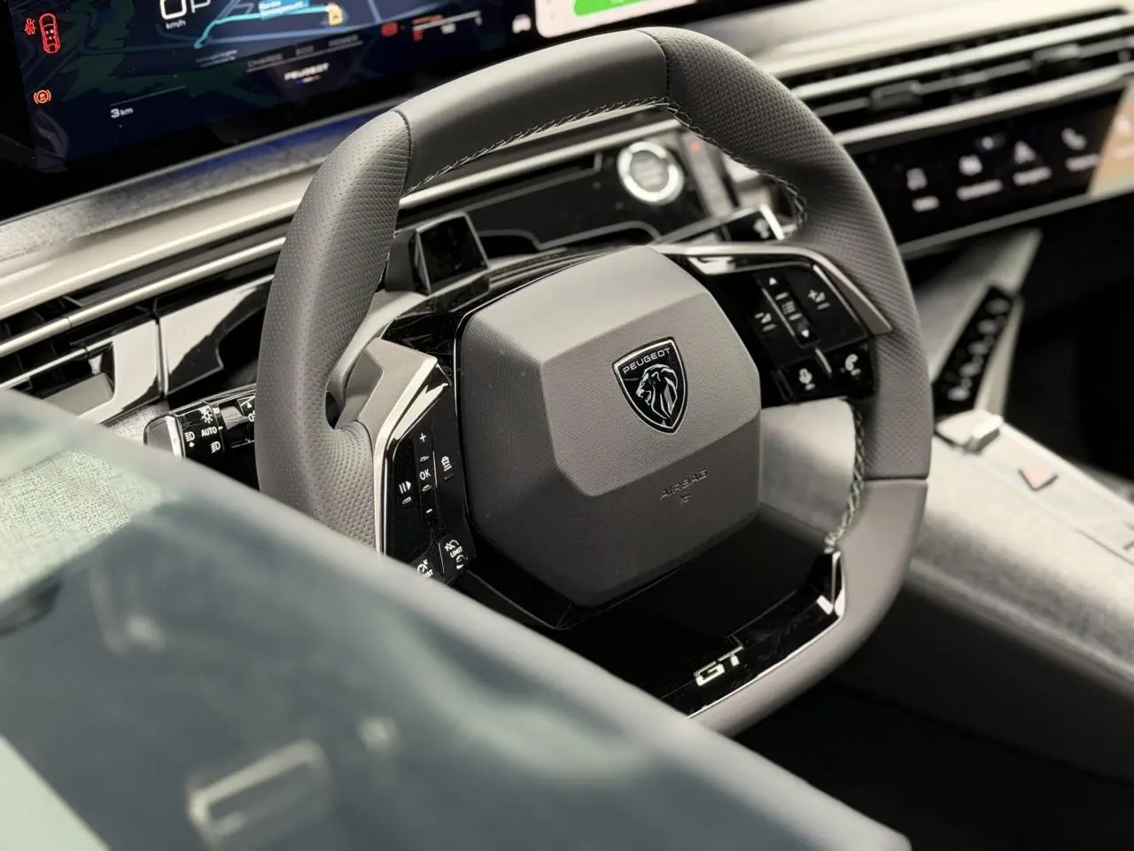 Gros plan sur le volant compact cuir perforé noir avec logo Peugeot et badge GT, tableau de bord numérique en arrière-plan.