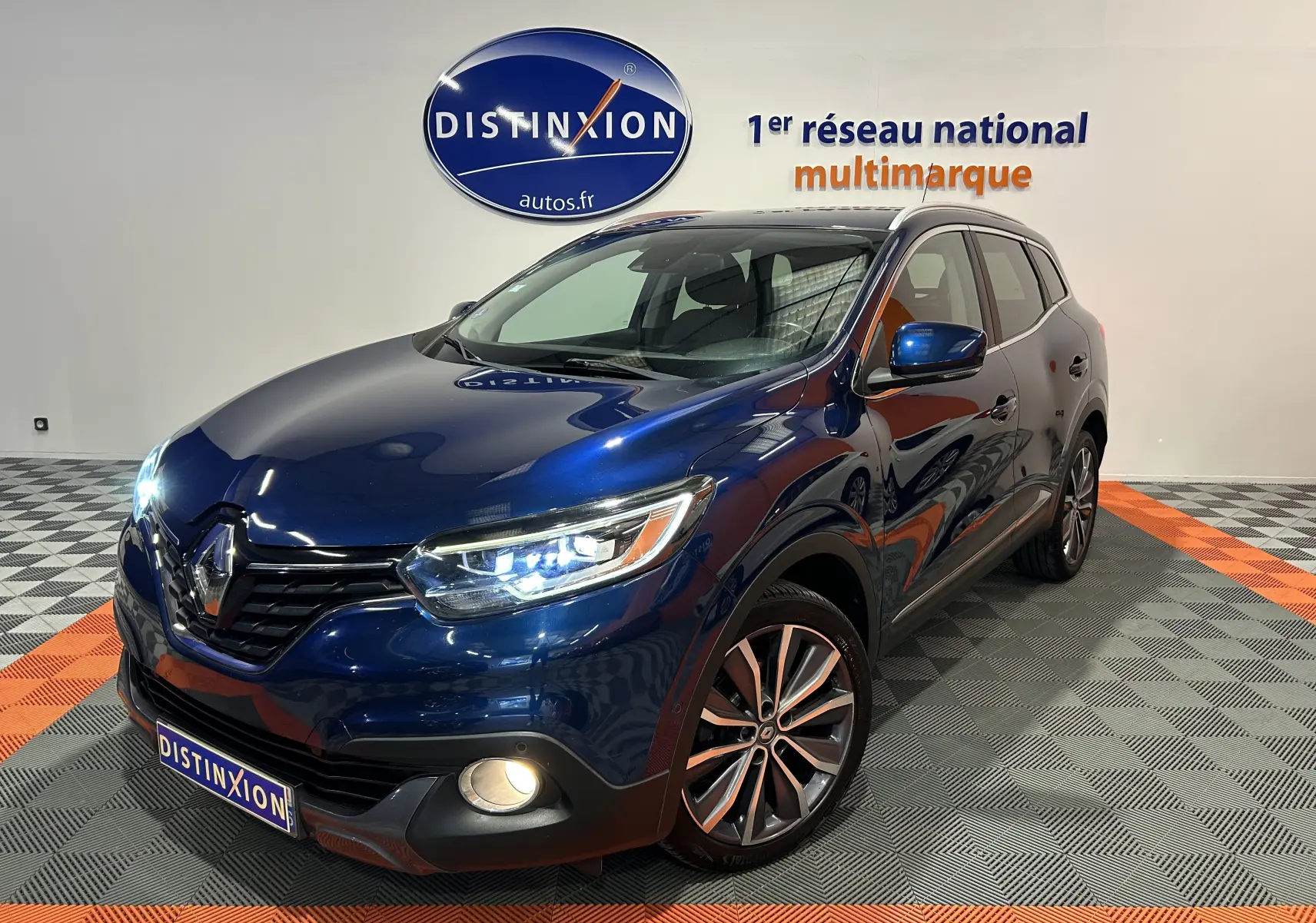Renault Kadjar TCe 130 Intens bleu en 3/4 avant droit avec phares allumés dans showroom multimarque.