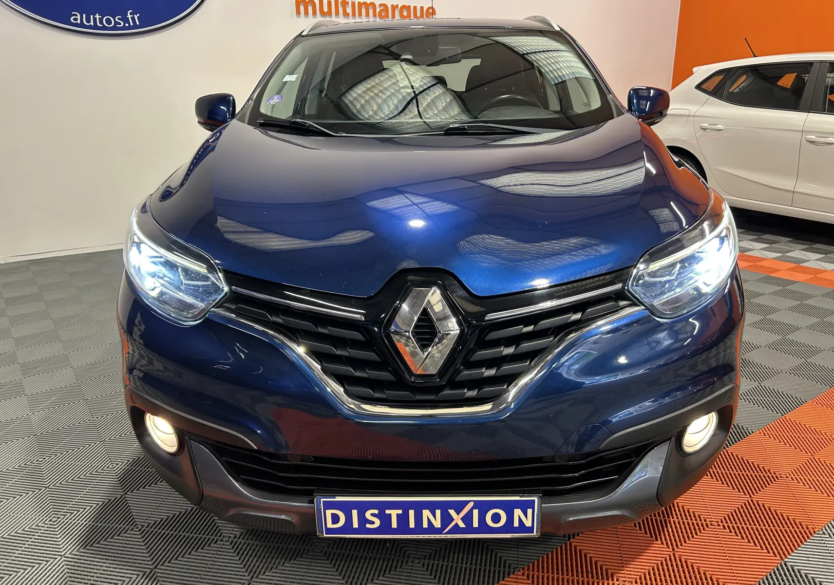 Vue avant rapprochée d'un Renault Kadjar bleu 2017 avec phares allumés et calandre chromée visible en intérieur showroom.