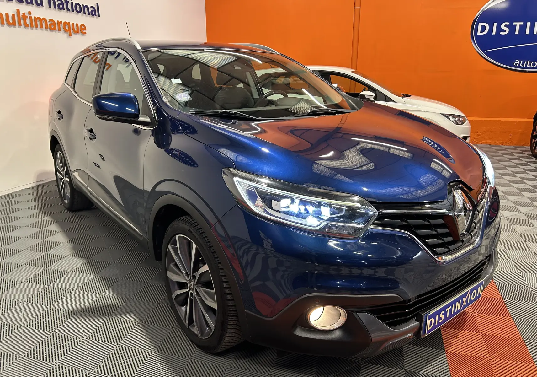 Renault Kadjar bleu en 3/4 avant droit avec phares LED allumés dans un showroom orange.