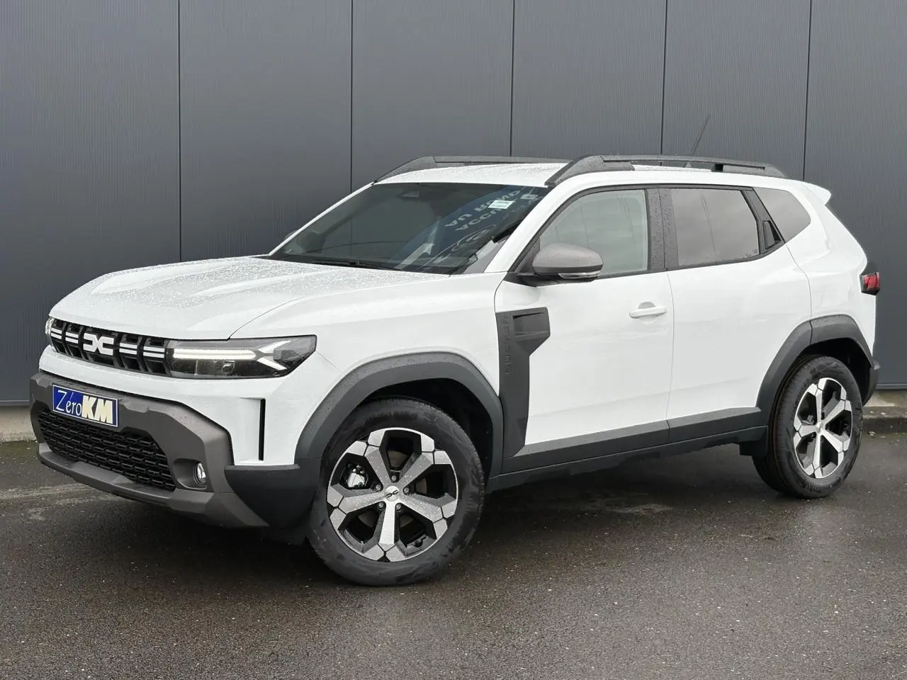 SUV Dacia Duster blanc glacier vu en 3/4 avant droit, avec jantes alliage et barres de toit noires.