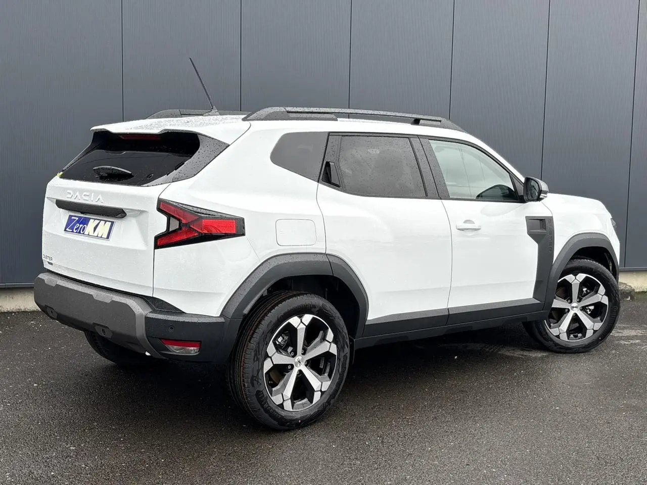 SUV Dacia Duster blanc glacier vu en 3/4 arrière droit, avec jantes alliage et barres de toit noires.