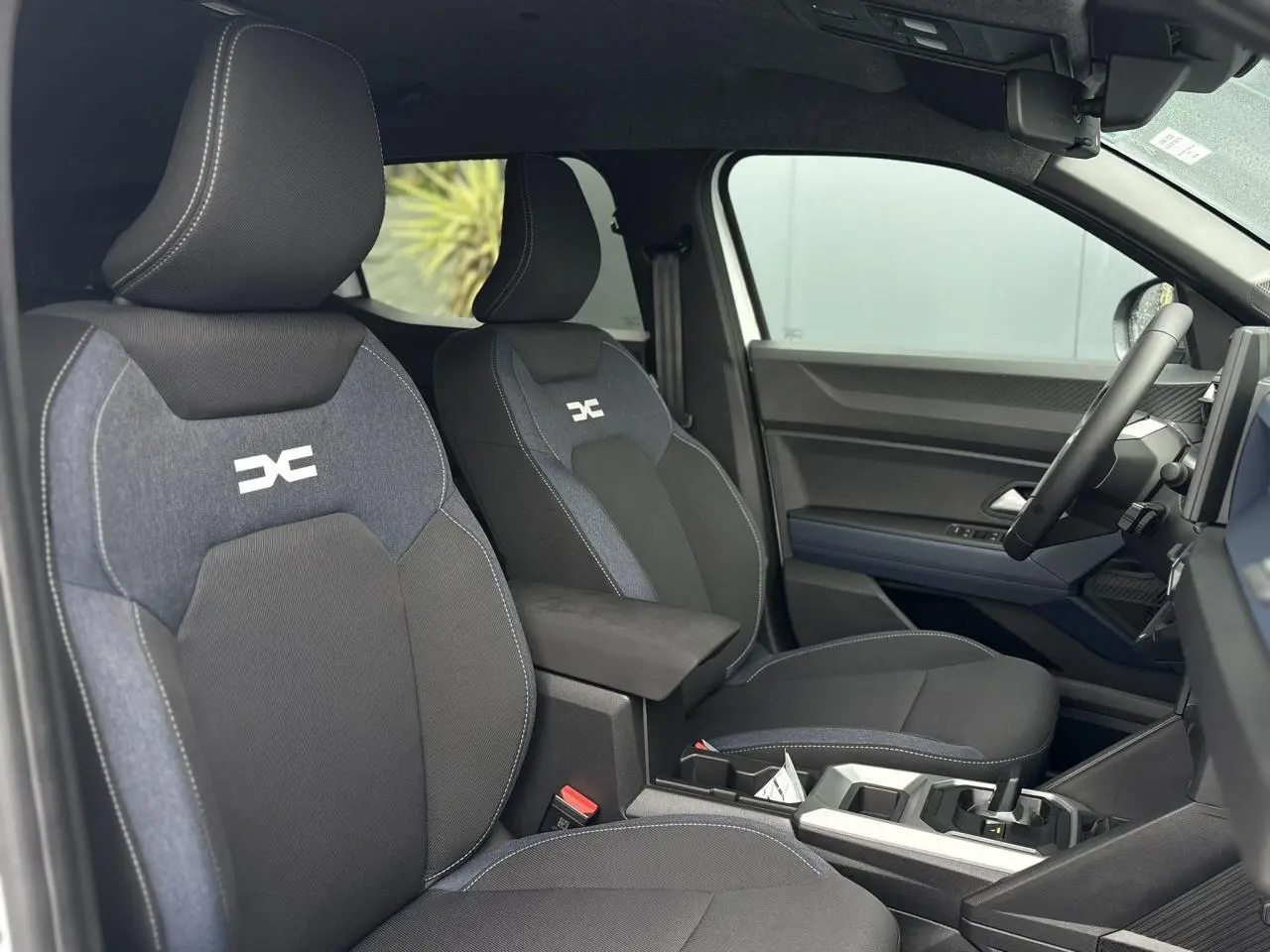Intérieur côté conducteur du Dacia Duster blanc Glacier 2025, sièges noirs avec logo DC et accoudoir central.