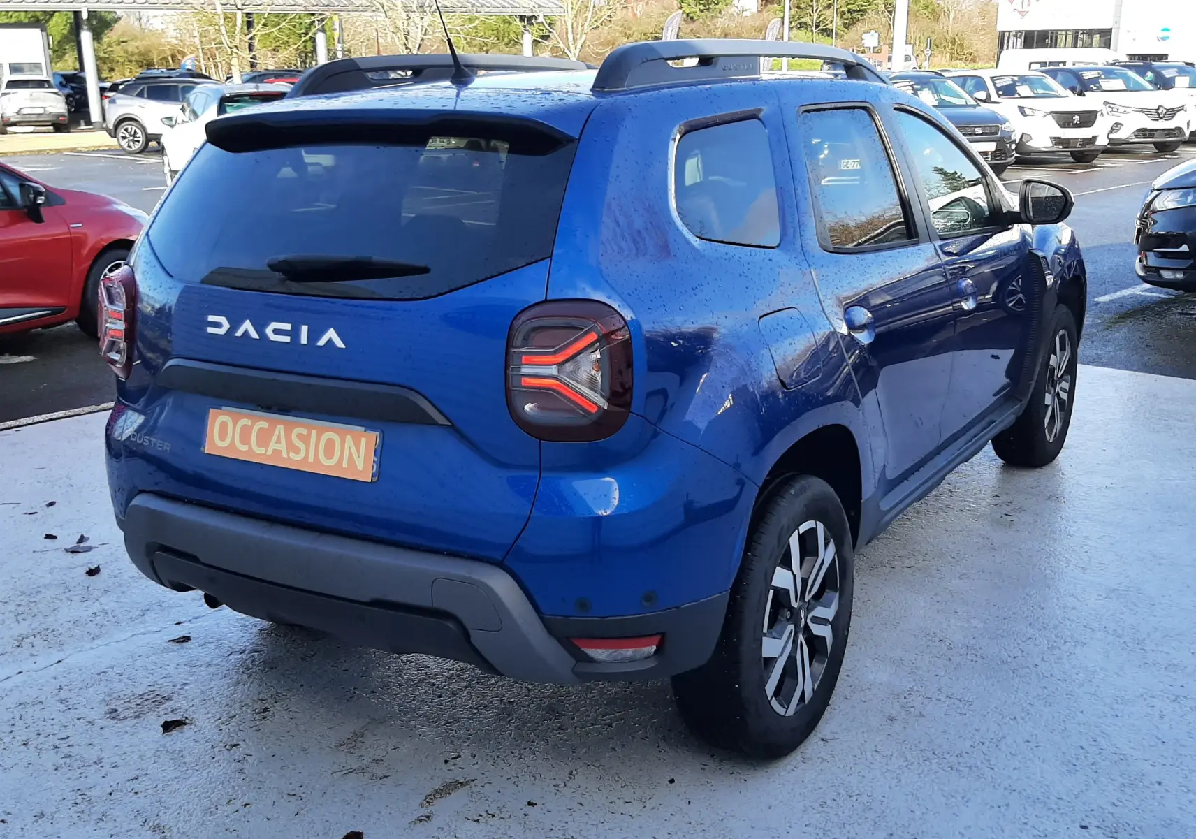 Vue 3/4 arrière droite d'un Dacia Duster bleu 2023 avec feux arrière LED et barres de toit noires sur parking.