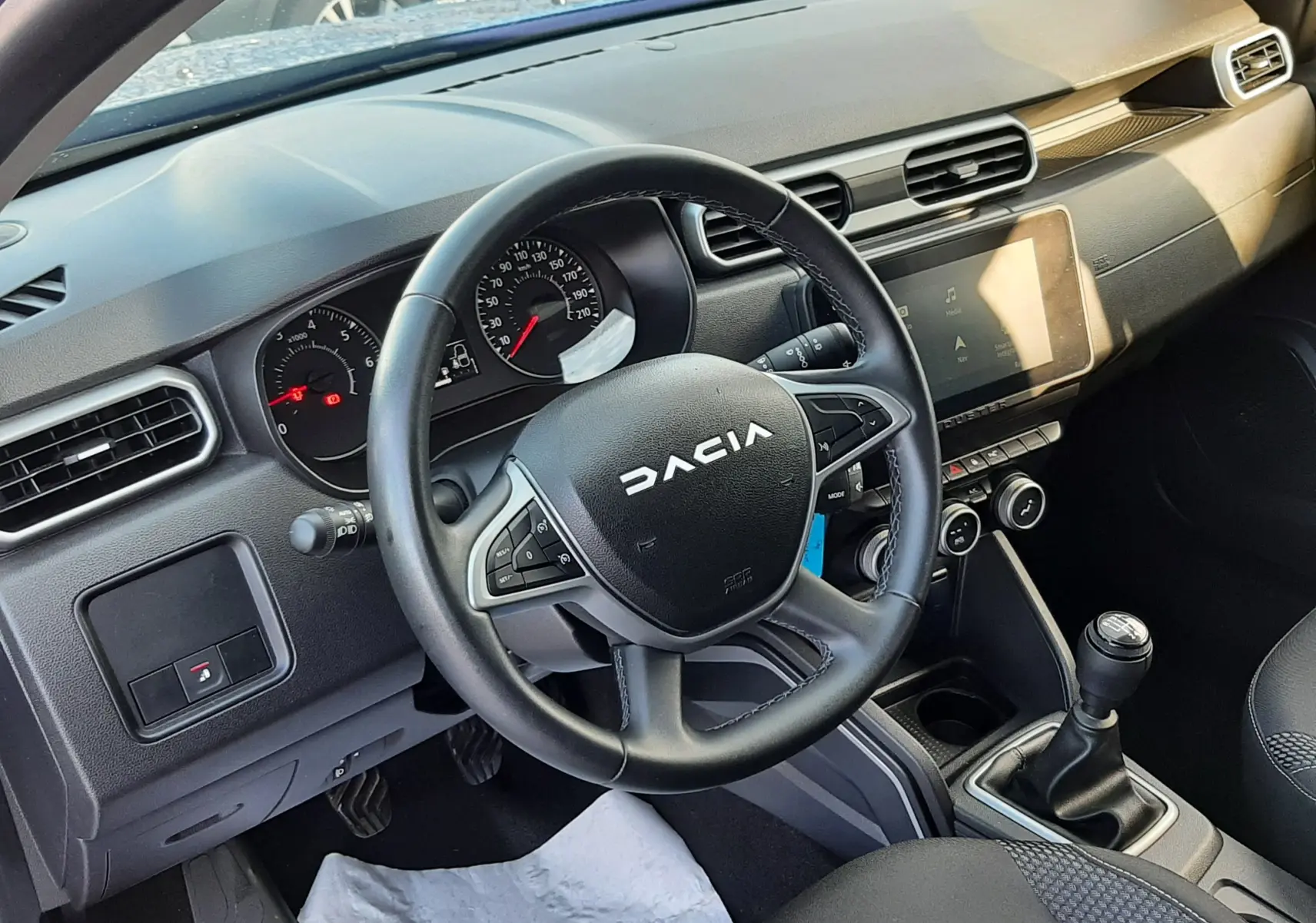 Vue intérieure côté conducteur du Dacia Duster 2023, volant multifonction, tableau de bord noir et levier de vitesses manuel.