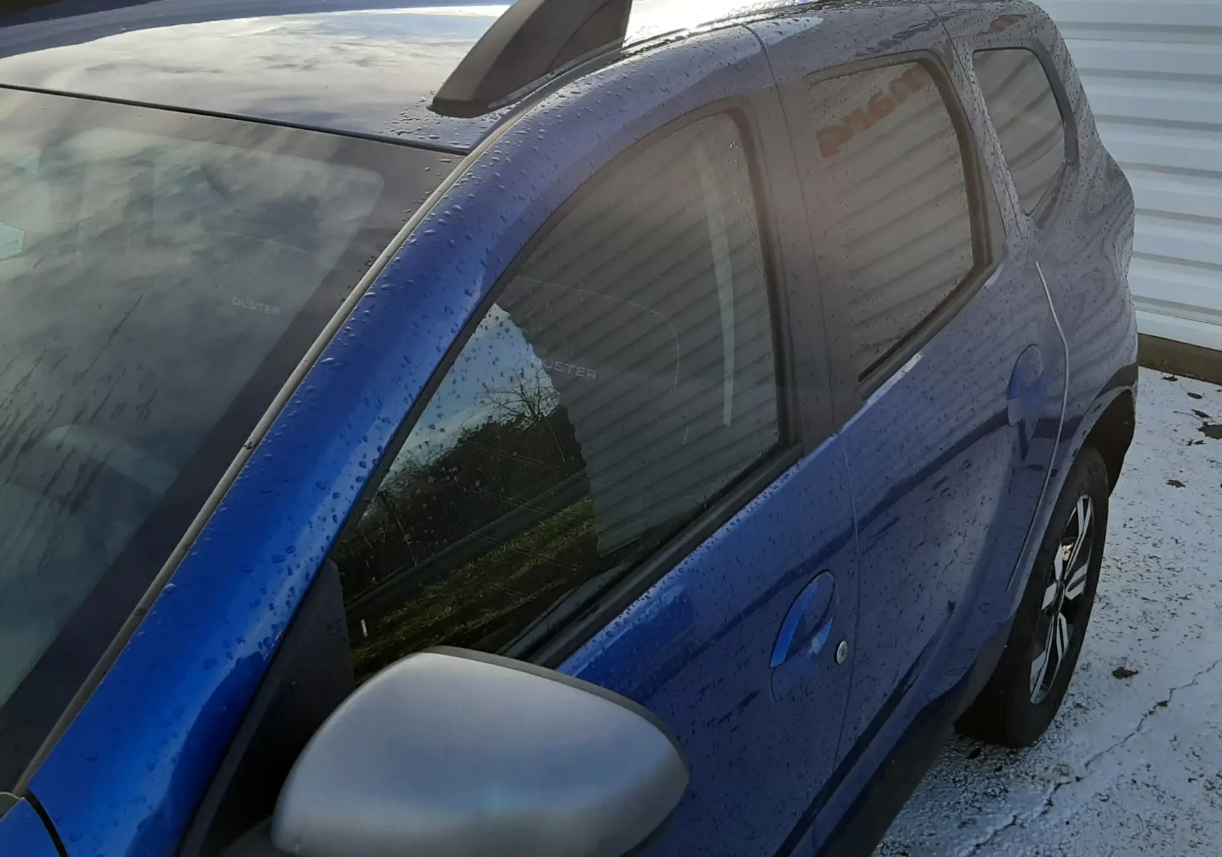 Vue 3/4 arrière droite d'un Dacia Duster bleu 2023 avec barres de toit noires et rétroviseur gris.