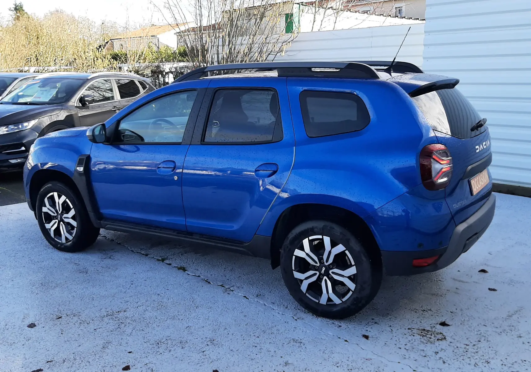 Vue 3/4 arrière droite d'un Dacia Duster bleu 2023 avec jantes bi-ton et barres de toit noires.