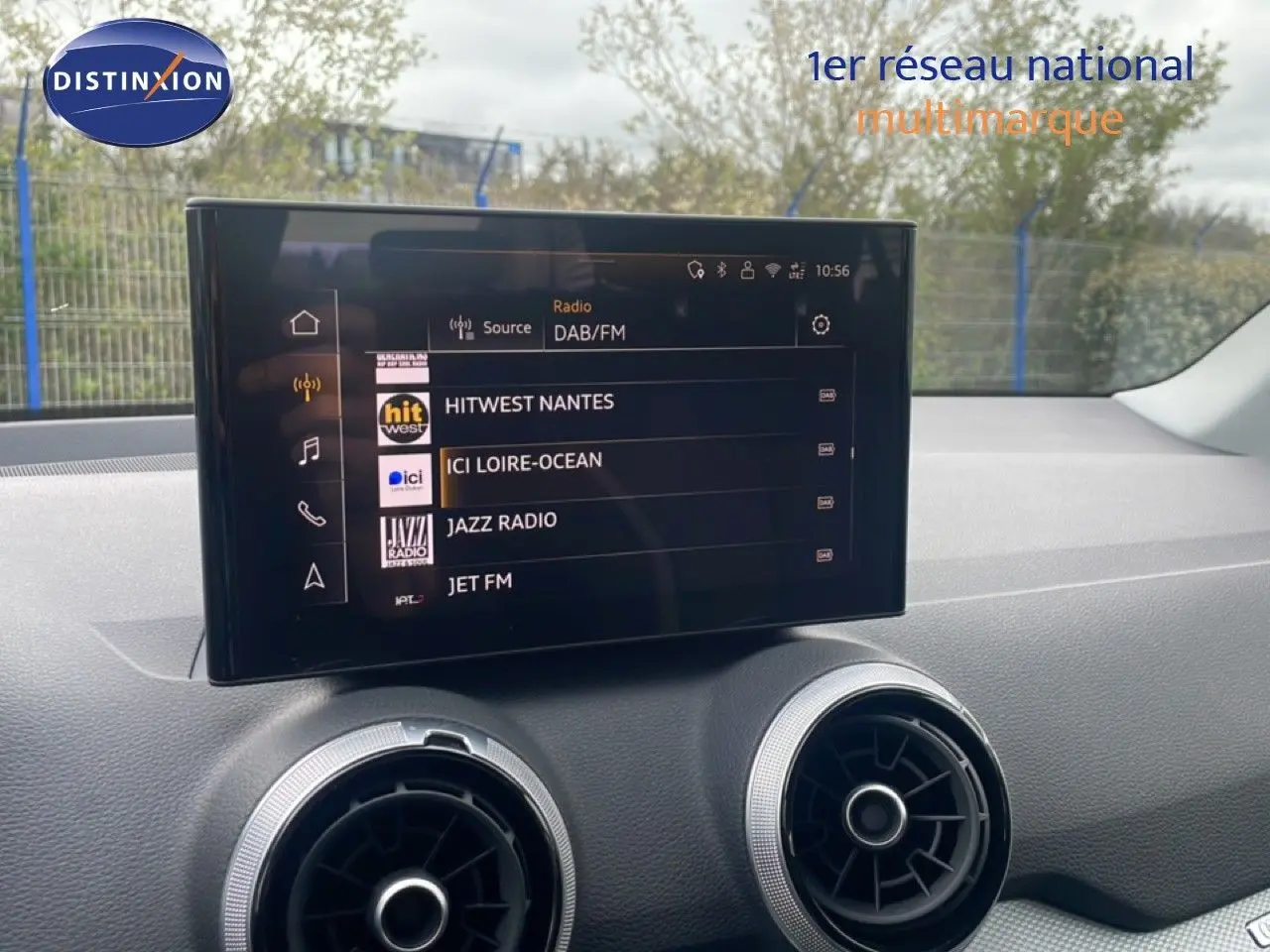 Écran tactile central affichant les stations radio, avec aérateurs ronds argentés sur tableau de bord noir.