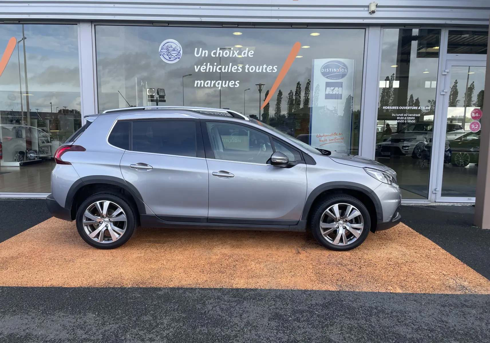 Profil droit d'un Peugeot 2008 gris 2019 garé devant une vitrine de concession avec jantes alliage visibles.