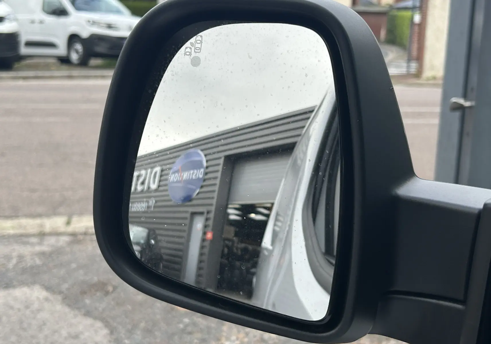 Rétroviseur latéral droit d'un Citroën Berlingo Fourgon blanc, reflétant un bâtiment commercial et un autre véhicule blanc.