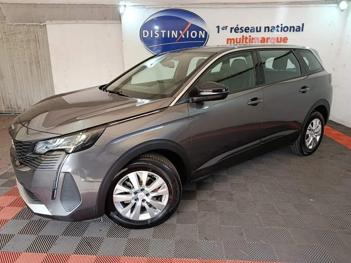 Peugeot 5008 gris clair vue 3/4 avant droit, avec jantes alliage et feux LED dans un showroom intérieur.