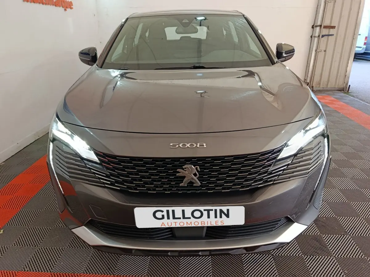Vue avant d'un Peugeot 5008 gris clair 2023 avec calandre noire et phares LED allumés dans un garage.
