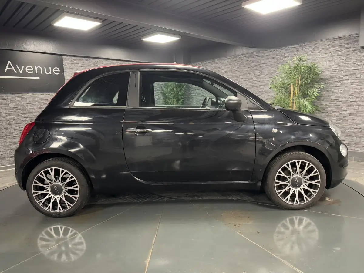 Profil côté gauche de la Fiat 500 noire avec jantes alliage et capote rouge visible en intérieur showroom.