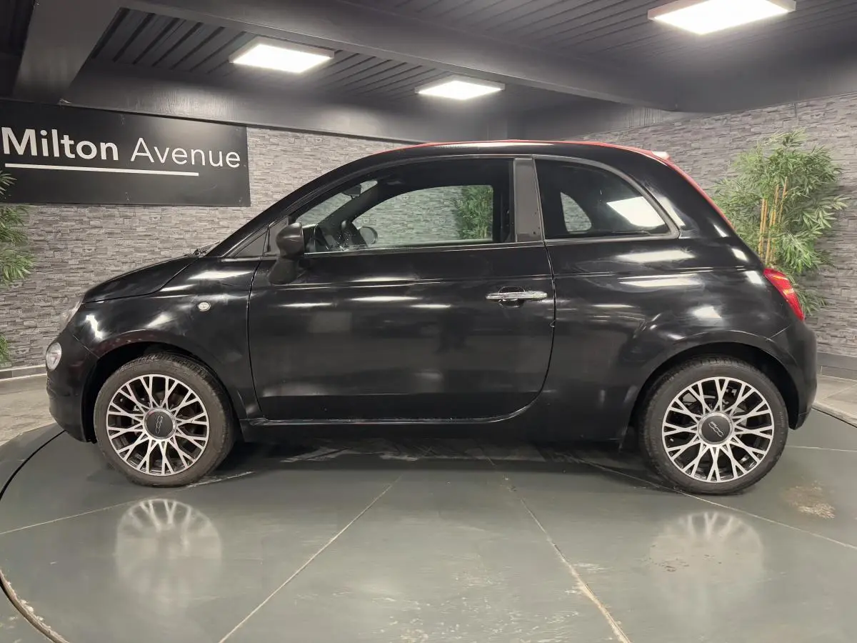 Profil côté gauche d'une Fiat 500 noire avec capote rouge et jantes alliage 16 pouces en intérieur showroom.