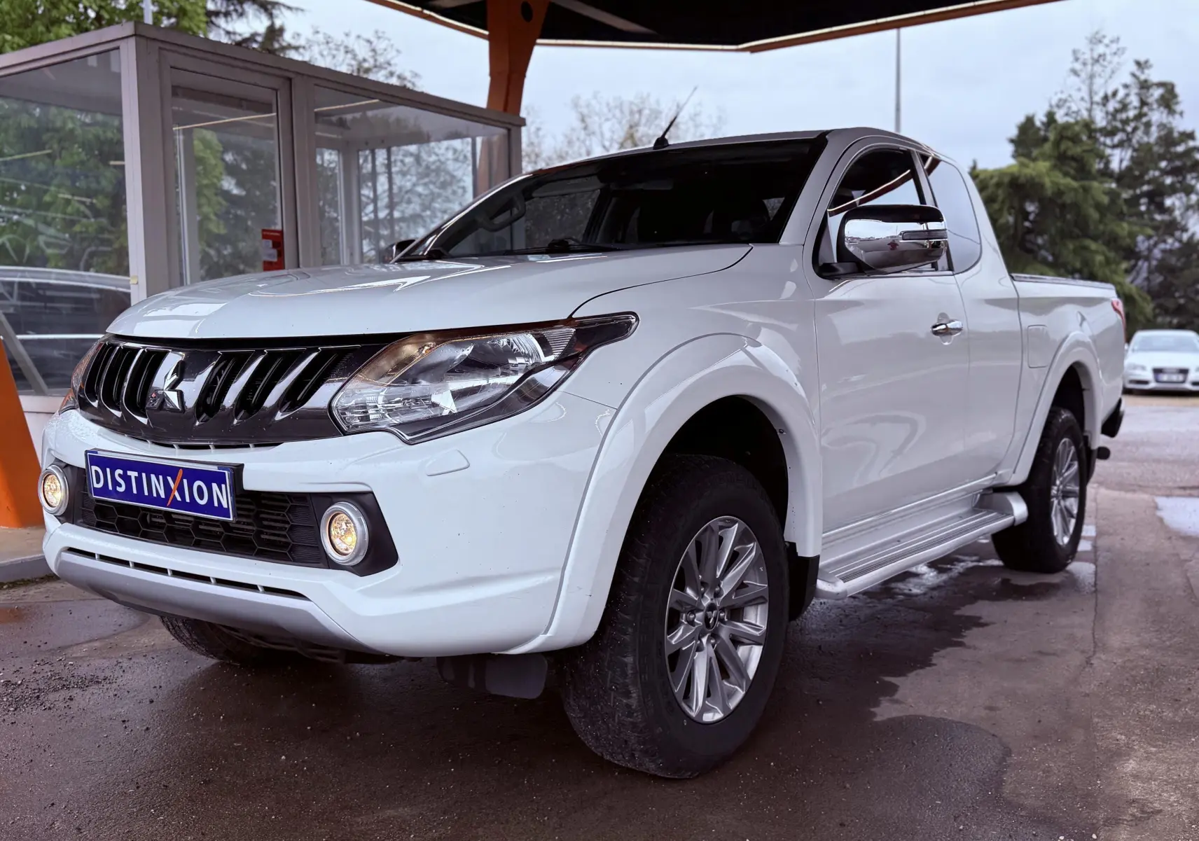 Mitsubishi L200 Club Cab blanc vu en 3/4 avant droit avec calandre noire et phares allumés sous abri.