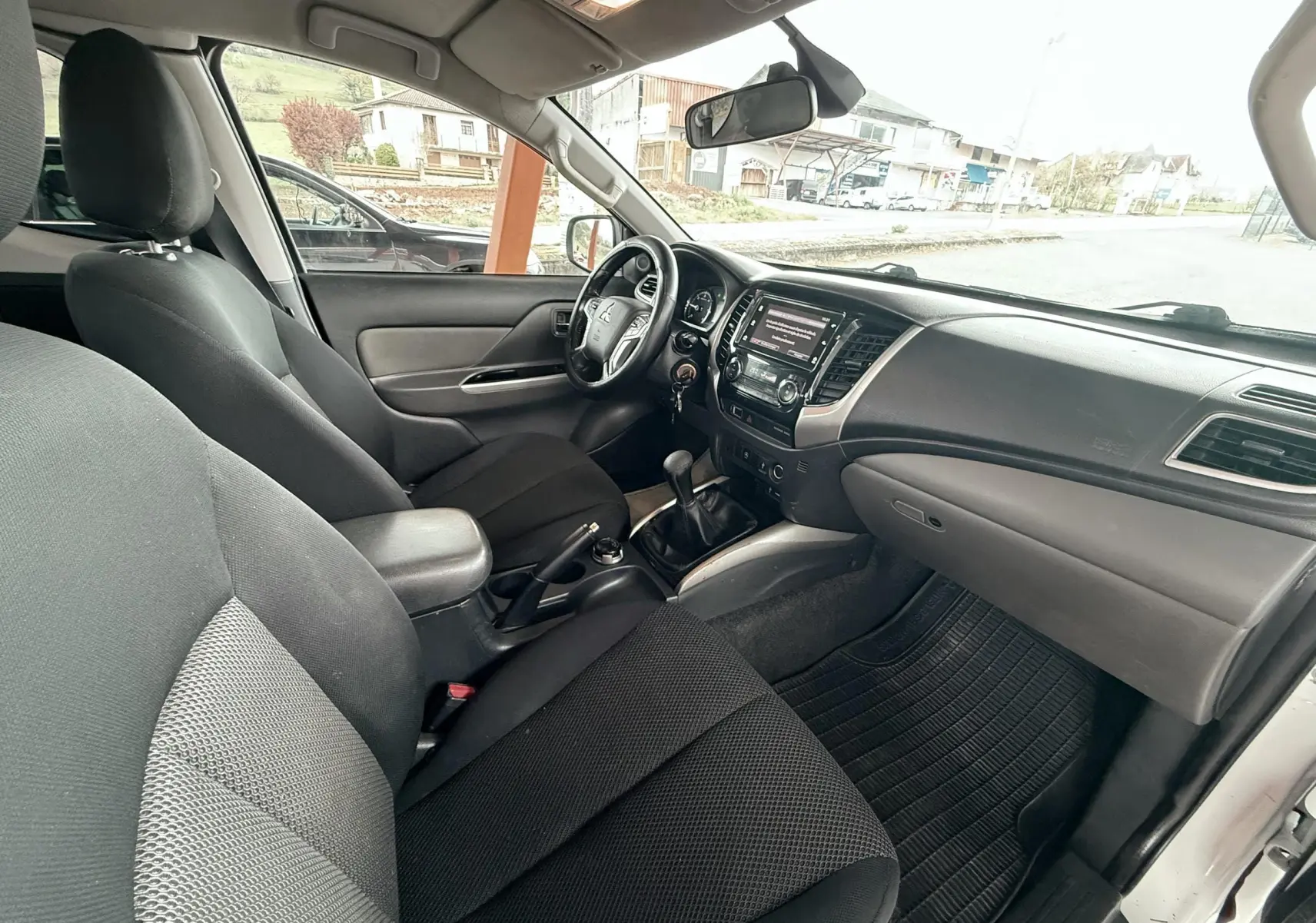 Vue intérieure côté conducteur du Mitsubishi L200 blanc, montrant le tableau de bord et les sièges noirs en tissu.