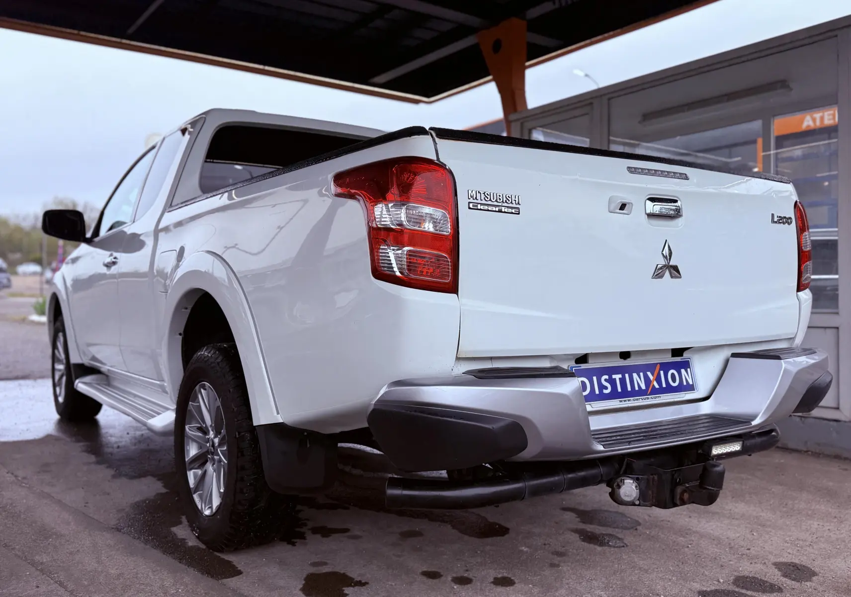 Vue 3/4 arrière droite du pick-up Mitsubishi L200 blanc avec hayon fermé et attelage visible sous le pare-chocs.