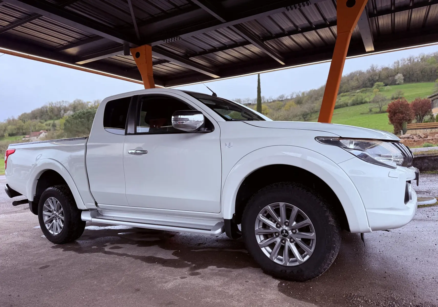 Vue 3/4 avant droit du pick-up Mitsubishi L200 Club Cab blanc sous un abri, avec jantes alliage et marchepieds visibles.