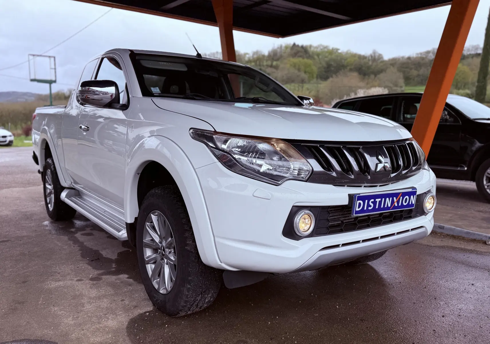 Vue 3/4 avant gauche d’un Mitsubishi L200 Club Cab blanc, avec calandre noire et antibrouillards allumés sous abri.