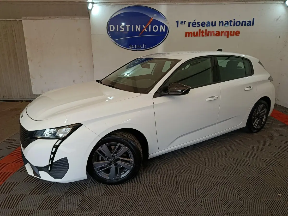 Peugeot 308 blanche vue en 3/4 avant droit, avec phares LED et jantes alliage foncé dans un showroom.