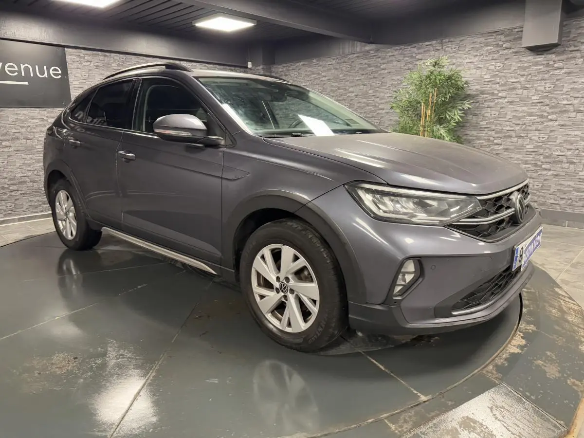 Volkswagen Taigo gris Ascot en 3/4 avant droit, jantes alliage Belmopan et feux LED allumés en intérieur showroom.