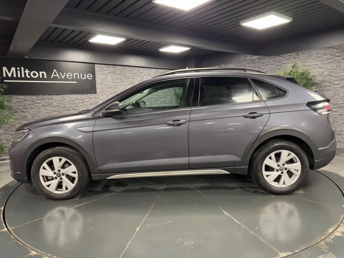 Profil droit d'un Volkswagen Taigo gris 2024 avec jantes alliage Belmopan et barres de toit noires dans un showroom.