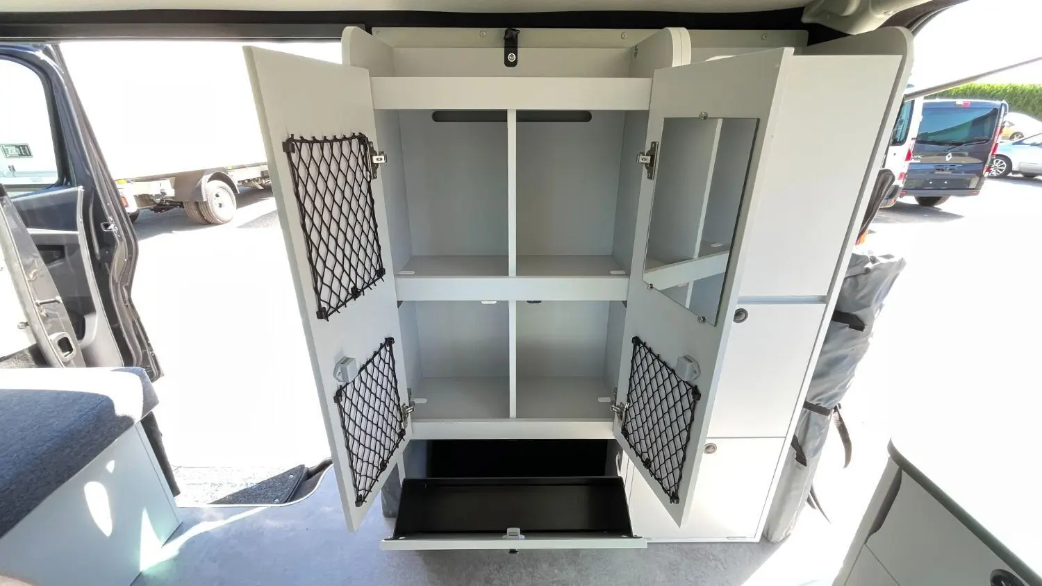 Vue intérieure du Renault Trafic Fourgon gris Comète montrant un meuble de rangement blanc ouvert avec filets et tiroir.