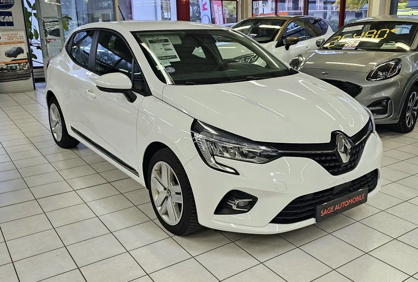 Renault Clio blanc glacier vue 3/4 avant droit en showroom, avec phares LED et calandre noire distinctive.