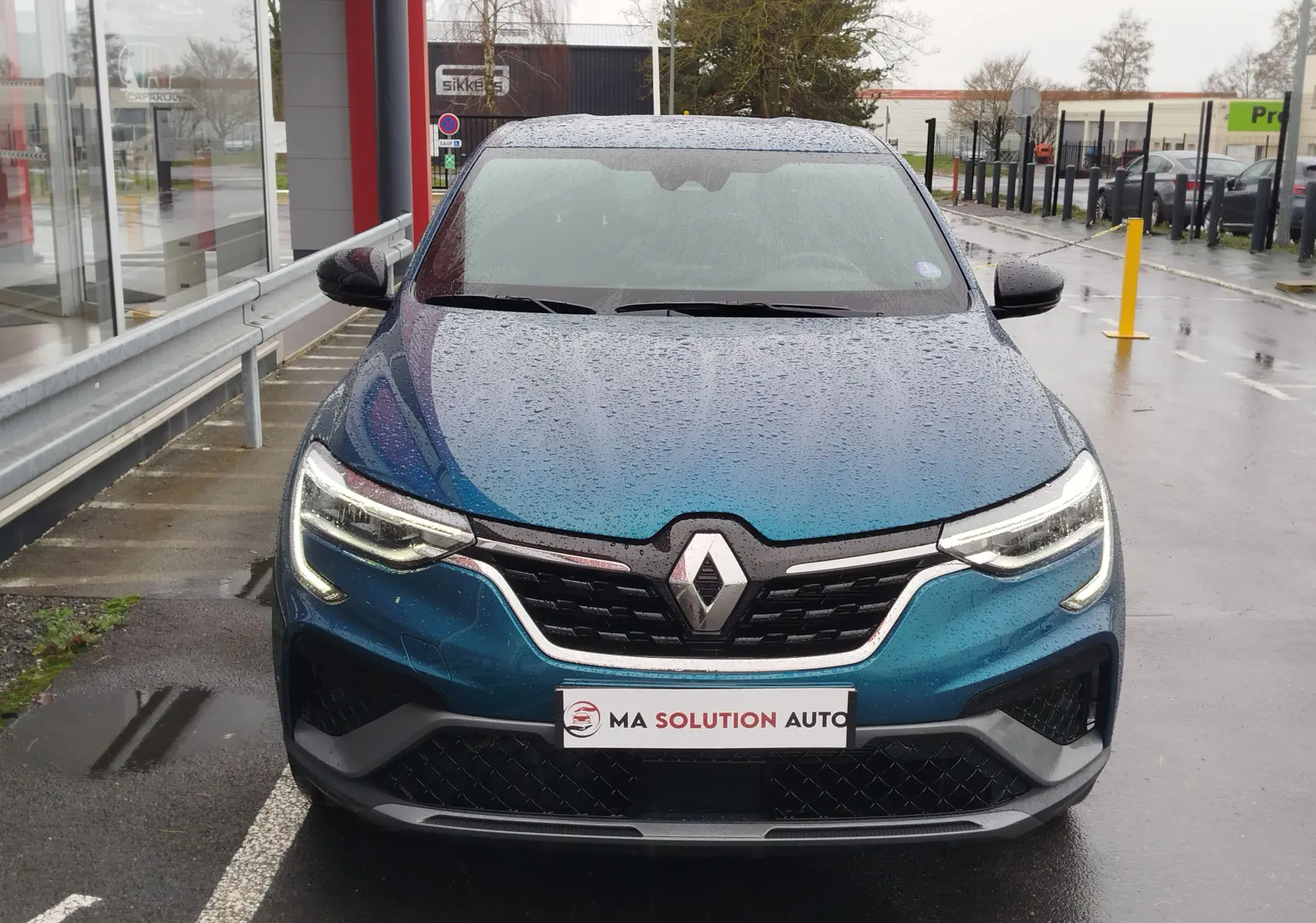 Vue frontale d'un Renault Arkana bleu Zanzibar 2022 sous la pluie, avec ses phares LED allumés et calandre noire distinctive.