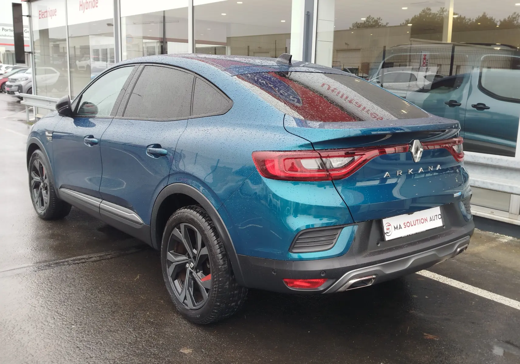 Vue 3/4 arrière droite d'un Renault Arkana bleu Zanzibar avec jantes noires et feux arrière LED sous la pluie.