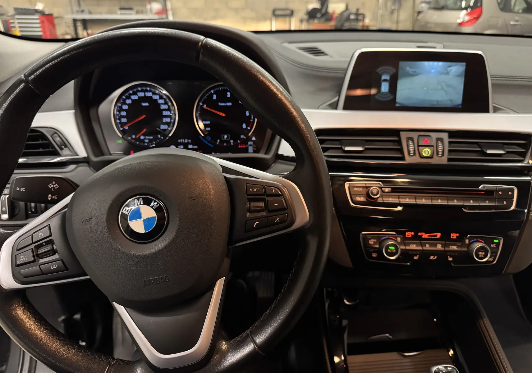 Vue intérieure centrée sur le volant cuir noir et tableau de bord avec écran caméra de recul d'une BMW X2 gris minéral 2019.