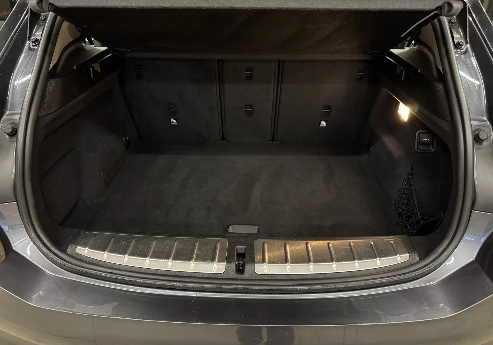 Coffre ouvert vu de l'arrière d'une BMW X2 gris minéral métallique, avec tapis de sol noir et éclairage intérieur.