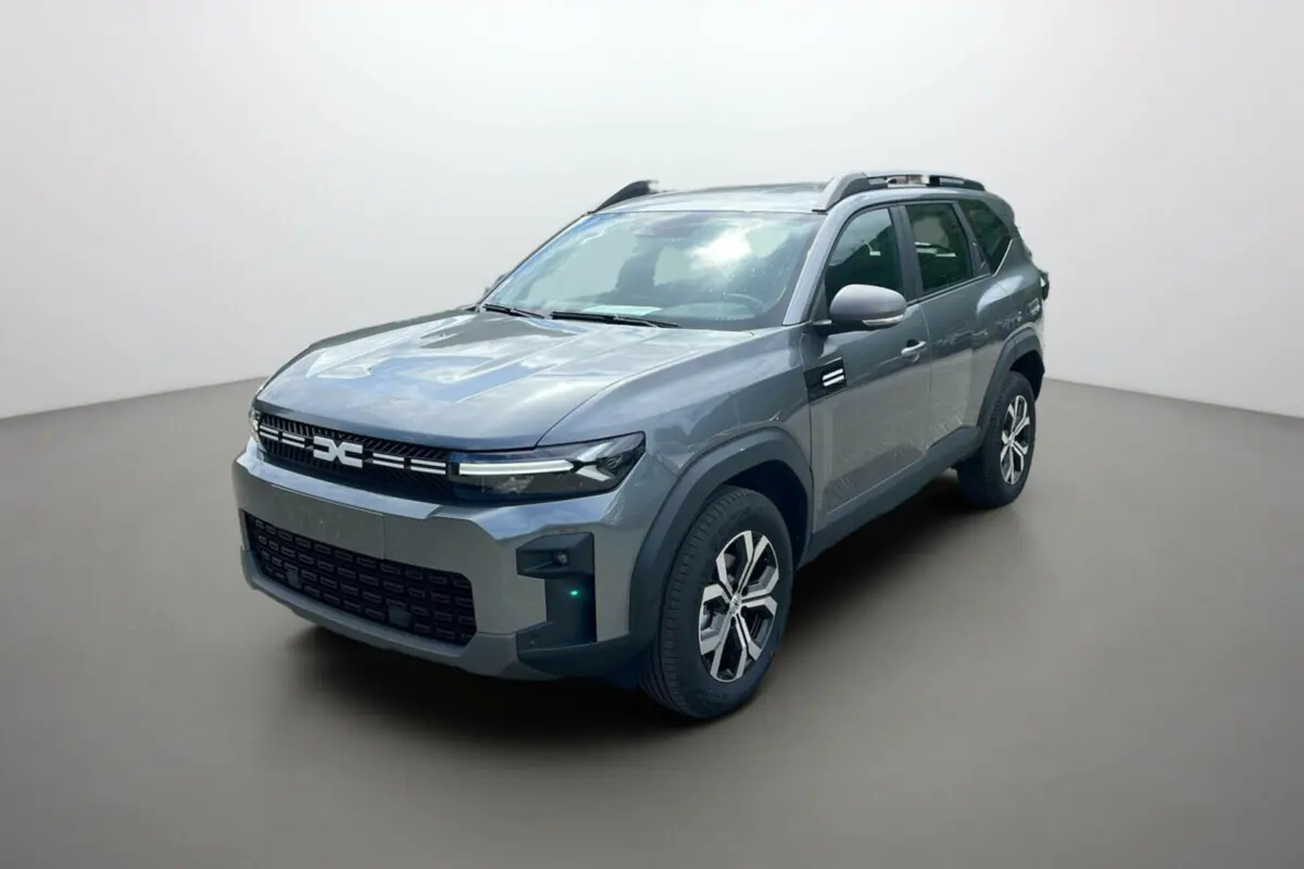 Dacia Bigster gris schiste en 3/4 avant droit, avec barres de toit et jantes alu 17 pouces visibles.