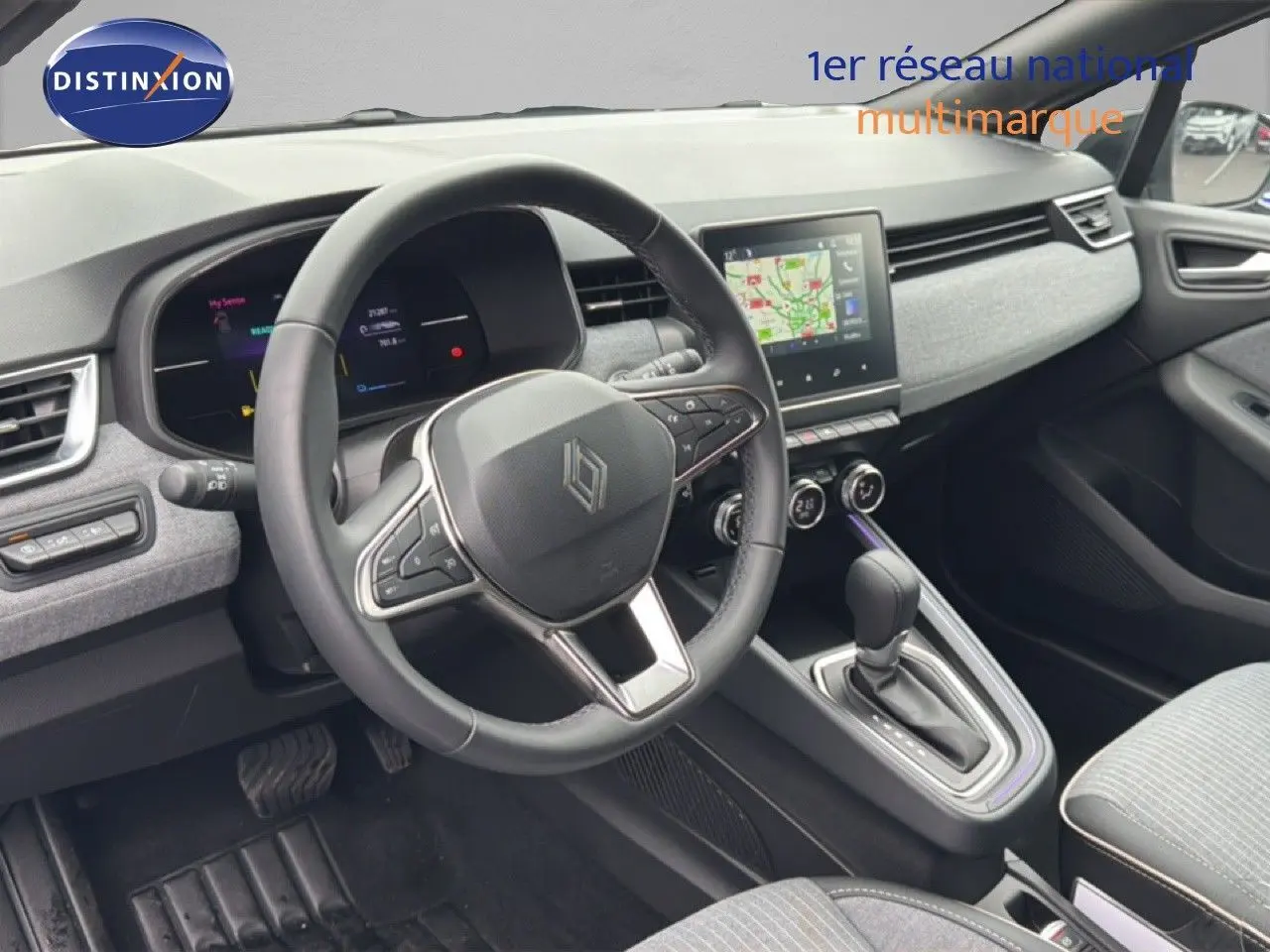 Vue intérieure côté conducteur de la Renault Clio 2025, tableau de bord gris clair avec écran tactile et volant cuir multifonction.