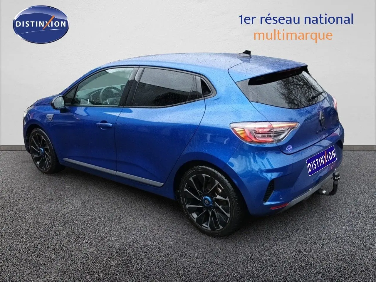 Vue 3/4 arrière gauche d'une Renault Clio 1.6 E-TECH hybride bleu Iron Metal avec jantes noires et attelage.